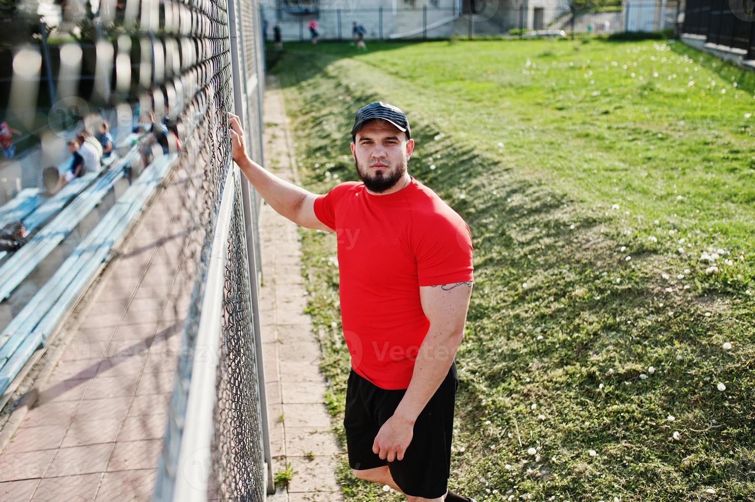 jeune homme musclé barbu brutal portant une chemise rouge, un short et une casquette au stade. photo
