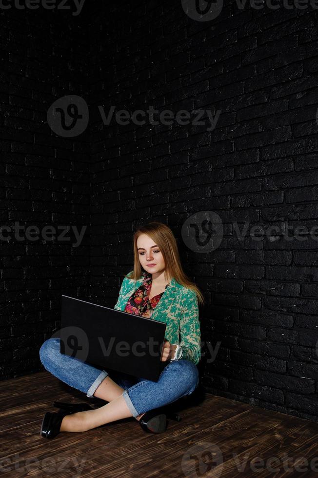 fille blonde élégante en veste et jeans avec ordinateur portable contre un mur noir en brique au studio. photo