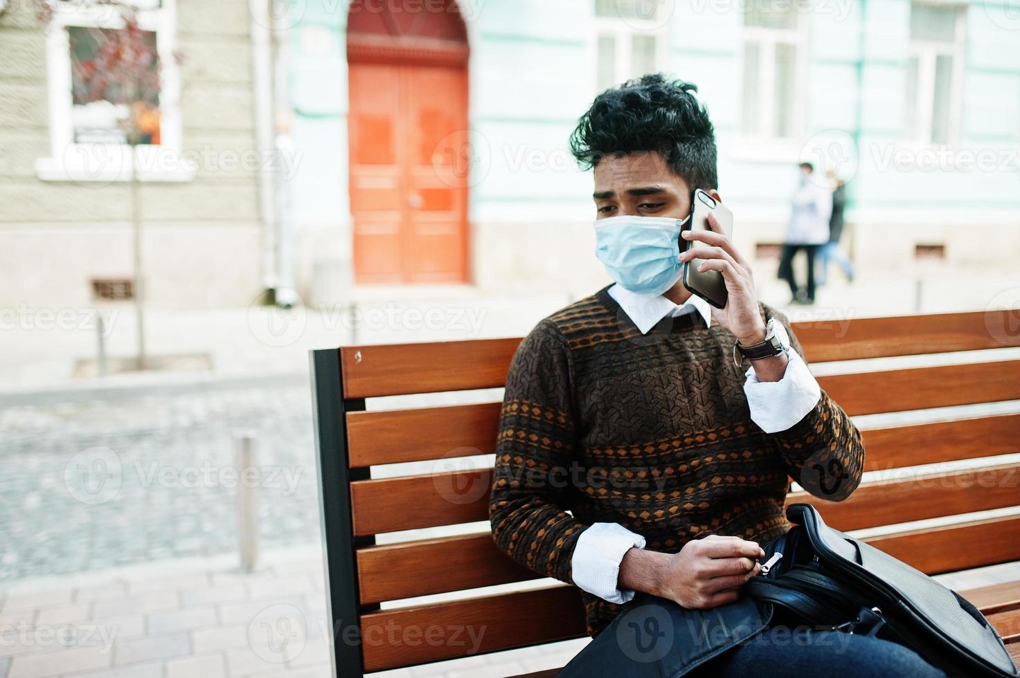 notion de coronavirus covid-19. homme indien d'asie du sud portant un masque pour se protéger du virus corona assis sur un banc avec un téléphone portable. photo