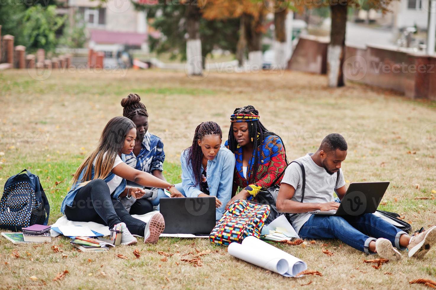 groupe de cinq étudiants africains qui passent du temps ensemble sur le campus de la cour universitaire. amis afro noirs assis sur l'herbe et étudiant avec des ordinateurs portables. photo