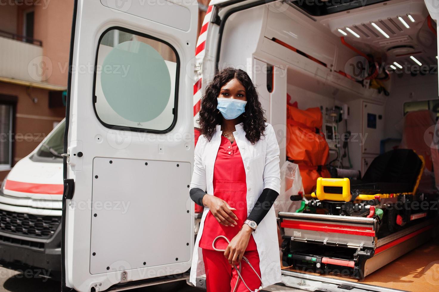 paramédic féminin afro-américain dans le masque médical de protection du visage debout devant la voiture d'ambulance. photo