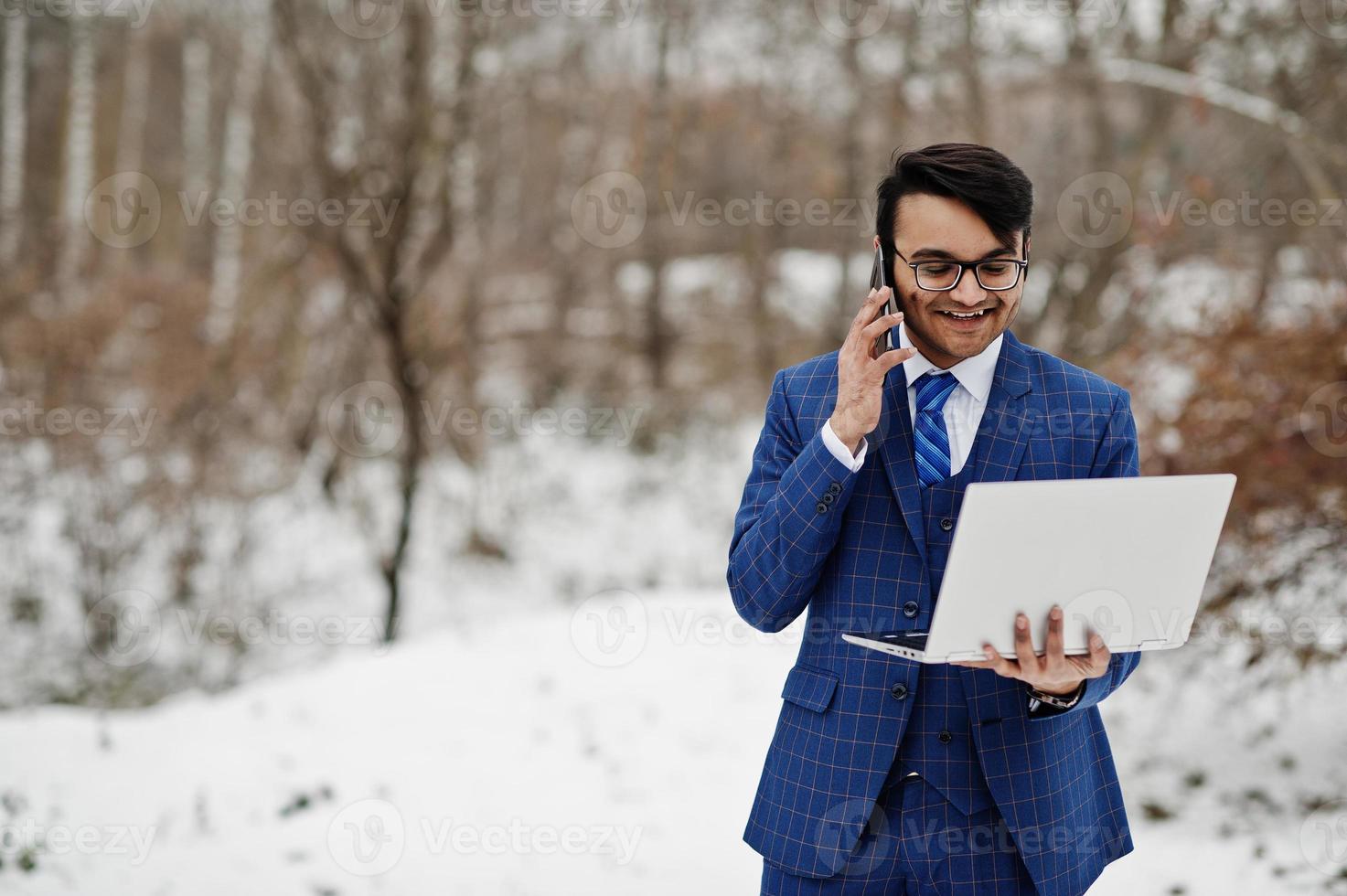 homme d'affaires indien élégant en costume et lunettes posé à la journée d'hiver en plein air avec un ordinateur portable à la main, parlant au téléphone mobile. photo