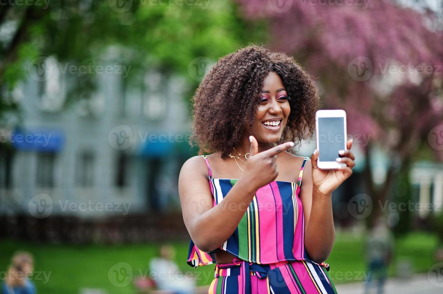 femme afro-américaine à la mode en combinaison rayée rose posée dans la rue de la floraison printanière et montre le doigt à l'écran du téléphone portable. photo