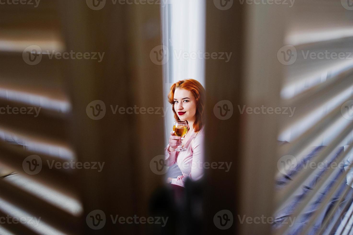 magnifique fille aux cheveux rouges en chemisier rose et jupe rouge avec un verre de vin à portée de main sur le lit de la chambre. photo