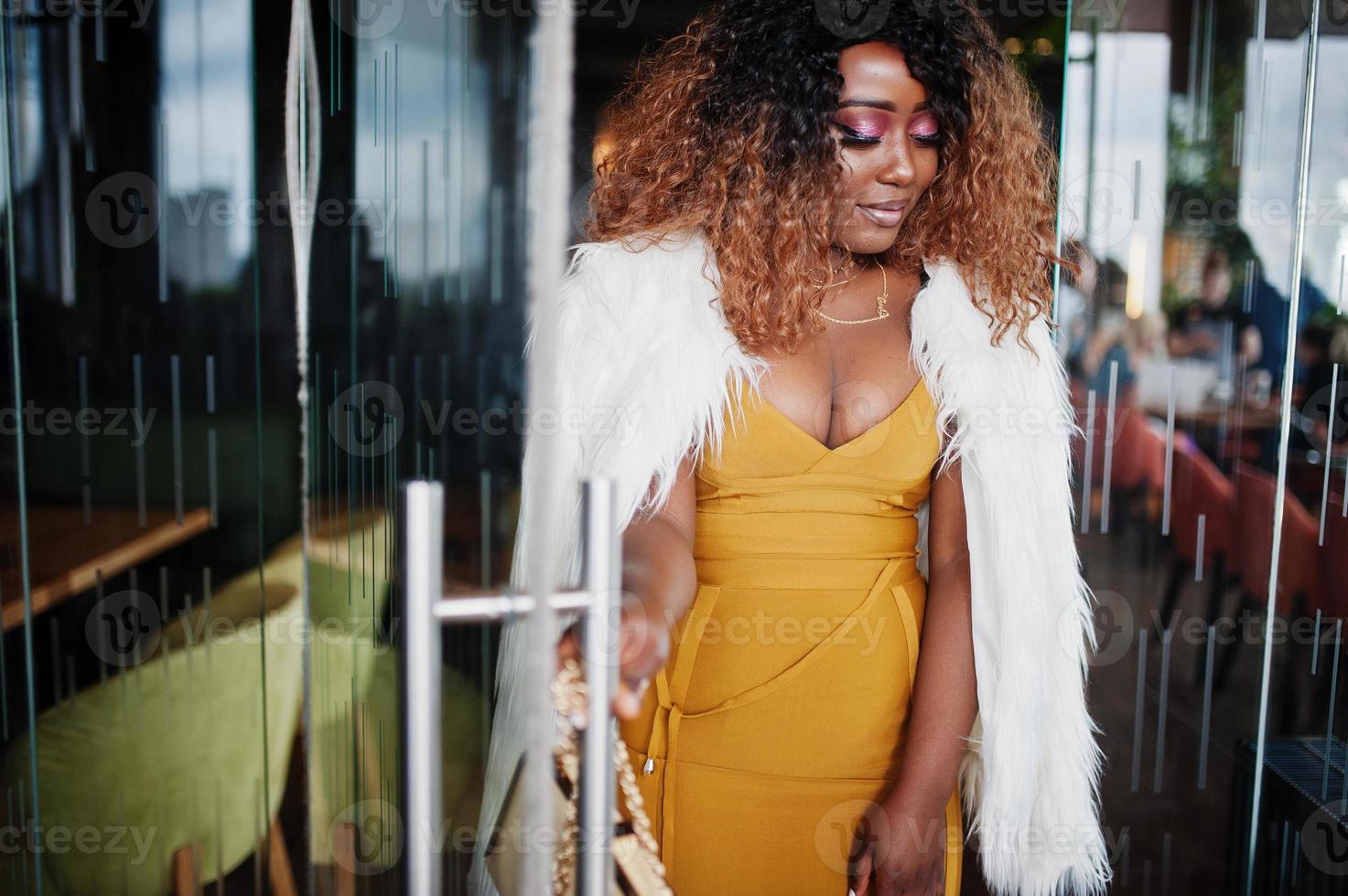 femme afro-américaine glamour en robe jaune et cape en laine blanche avec sac à main posé à l'entrée de la porte vitrée du restaurant. photo