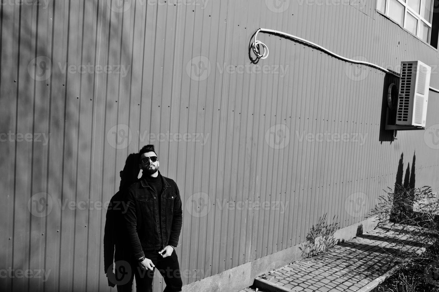 homme arabe de mode porter sur une veste en jean noir et des lunettes de soleil posées sur fond de mur en acier rouge. mec modèle arabe élégant et à la mode. photo