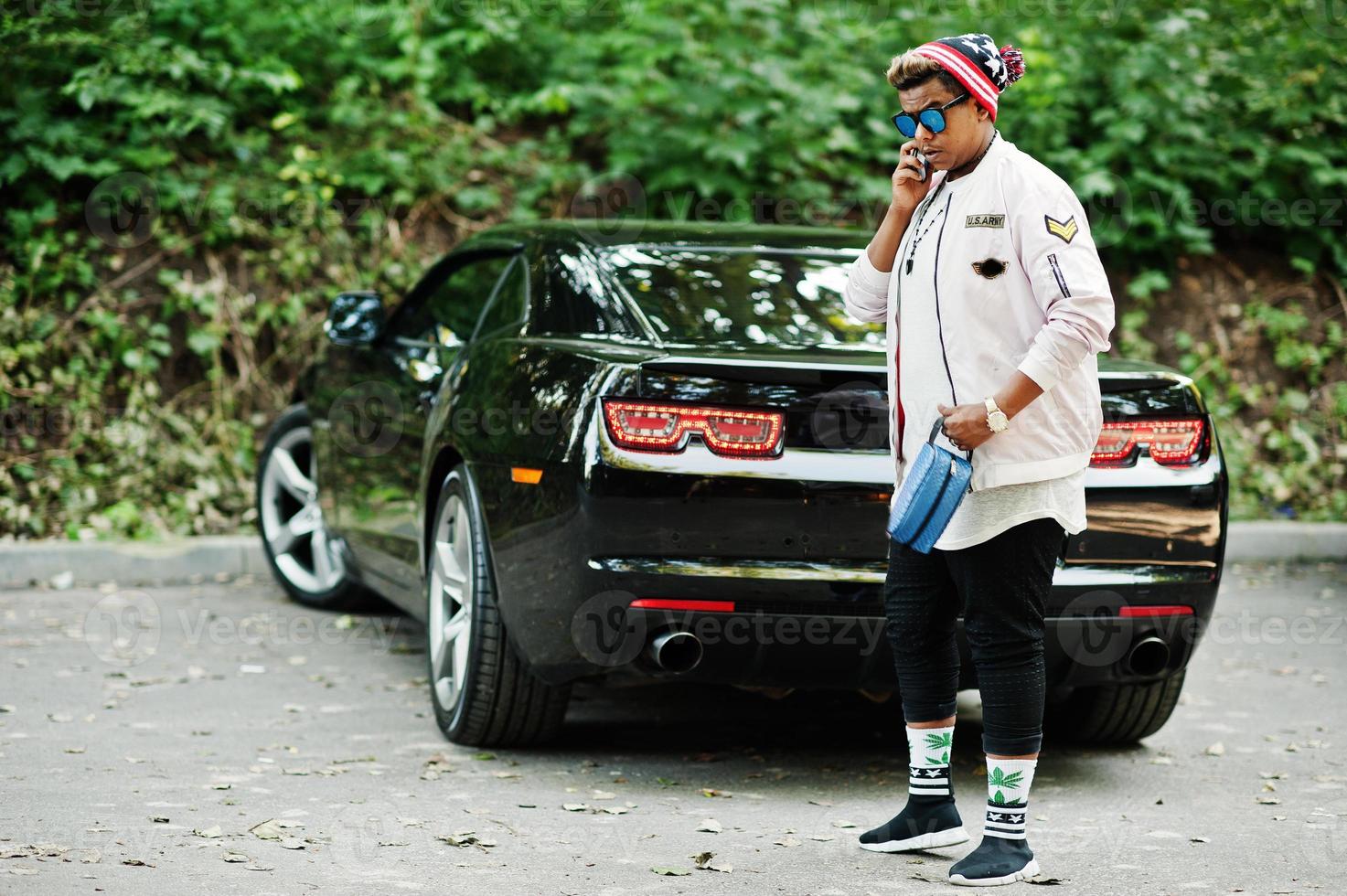 homme arabe hipster élégant avec chapeau et lunettes de soleil posés en plein air dans la rue contre sa voiture de muscle noire et parlant au téléphone mobile. photo