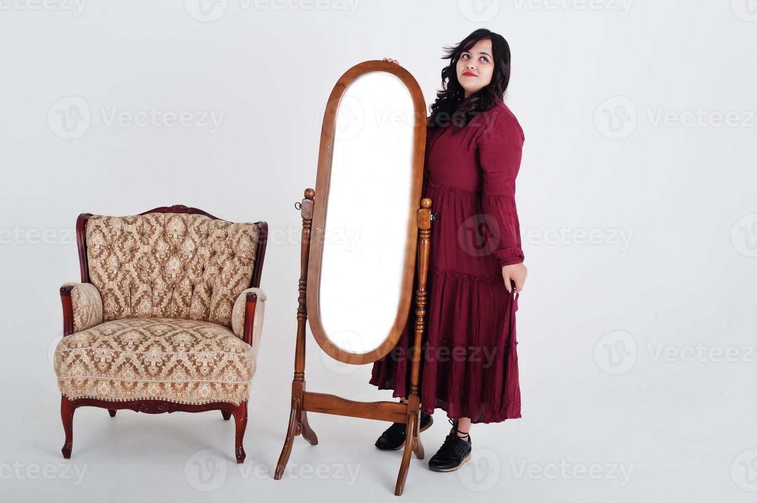 jolie femme sud-asiatique en robe rouge foncé posée au studio sur fond blanc contre le miroir et la chaise. photo