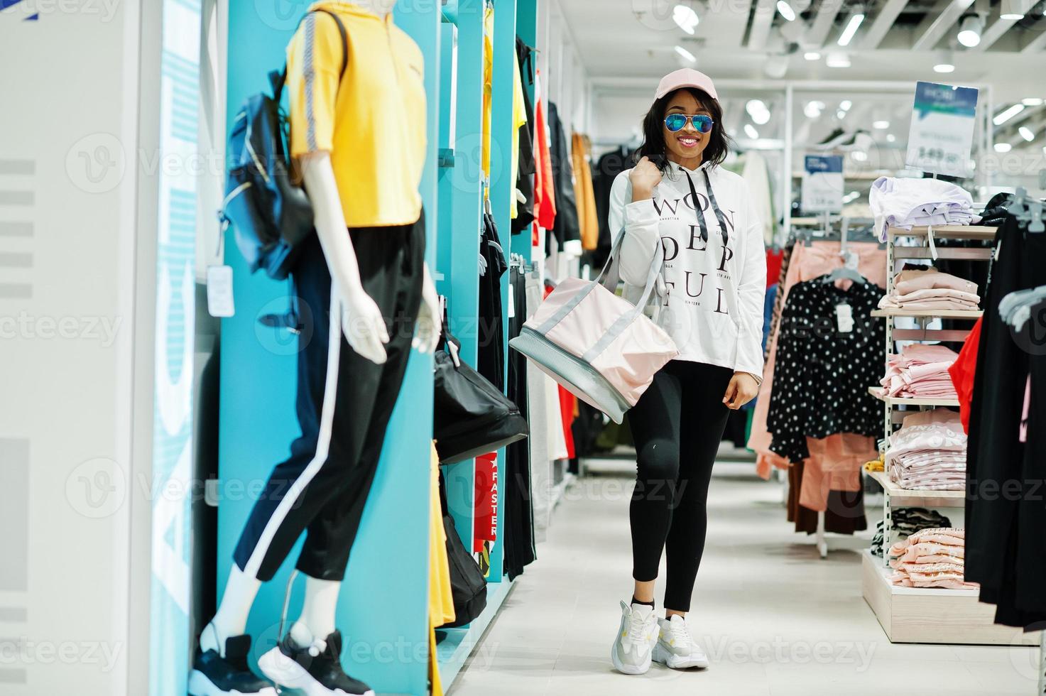 femmes afro-américaines en survêtements et lunettes de soleil shopping au centre commercial de vêtements de sport avec sac de sport contre les étagères. thème du magasin de sport. photo