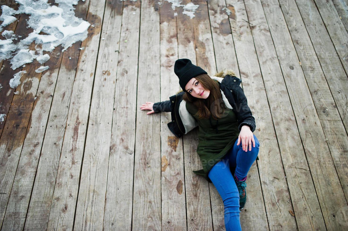 jeune fille porte un long sweat-shirt vert, un jean et un couvre-chef noir assis sur un plancher en bois avec de la neige. photo