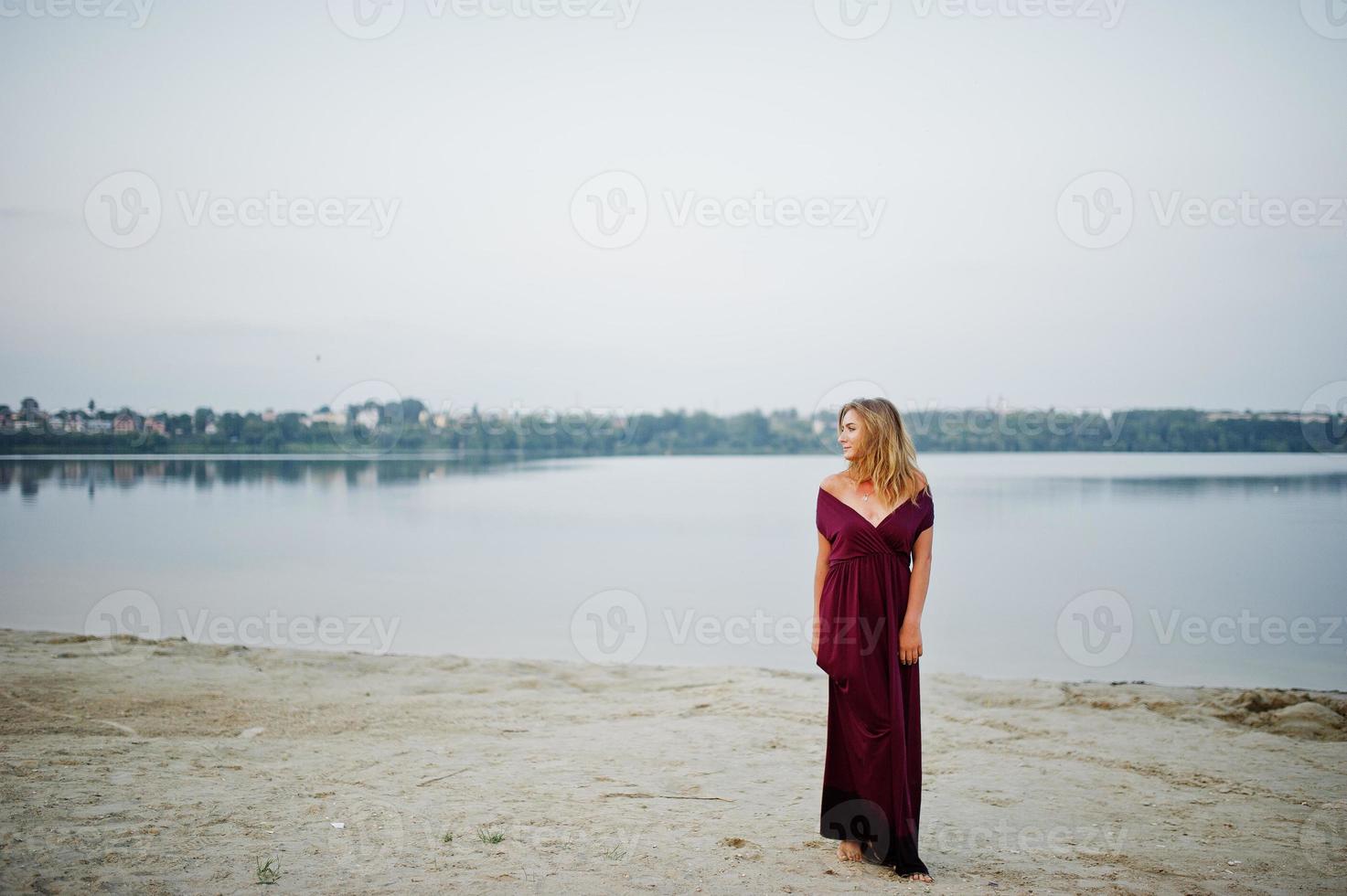 femme aux pieds nus sensuelle blonde en robe de marsala rouge posant contre le lac sur le sable. photo