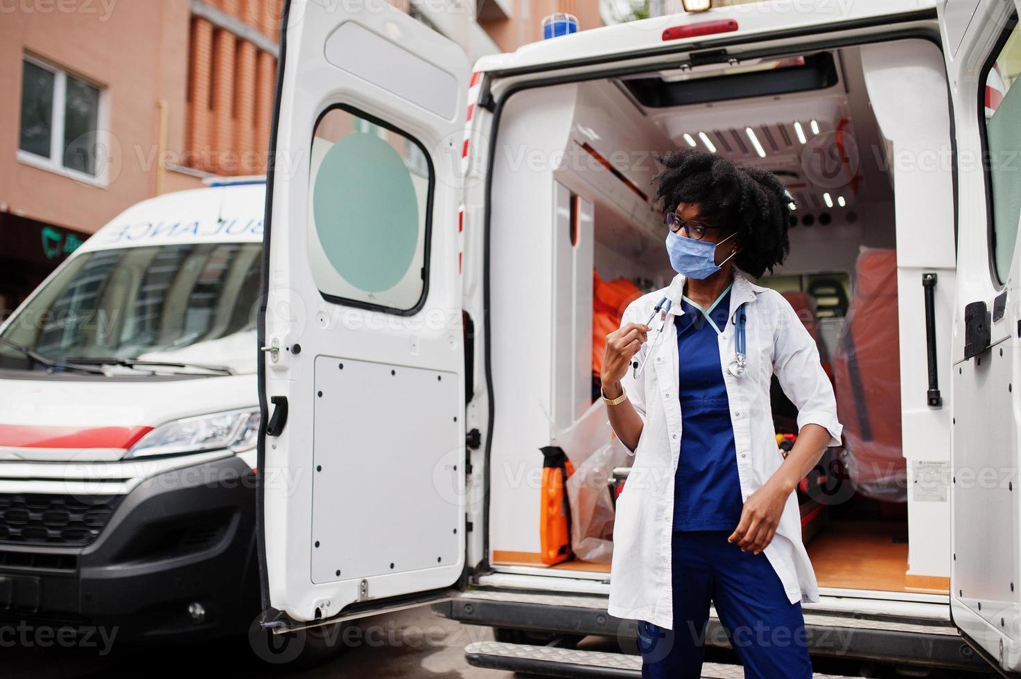 paramédic féminin afro-américain dans le masque médical de protection du visage debout devant la voiture d'ambulance. photo