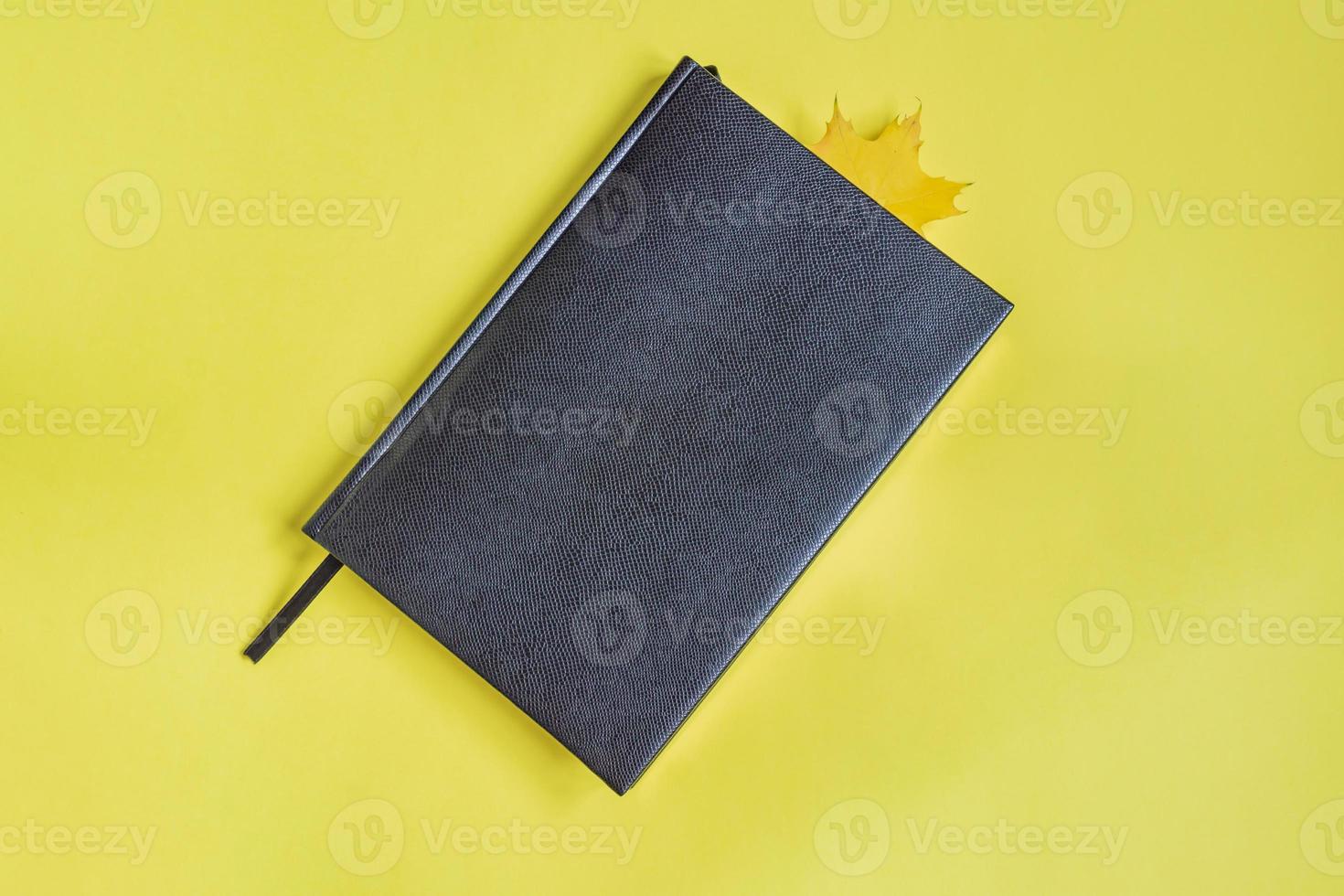 carnet en faux cuir de reptile de couleur noire avec feuille d'érable comme signet sur fond jaune. mise à plat minimale photo