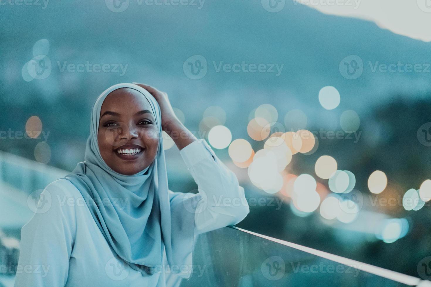 femme musulmane africaine dans la nuit sur un balcon souriant à la caméra avec des lumières bokeh de la ville en arrière-plan. photo