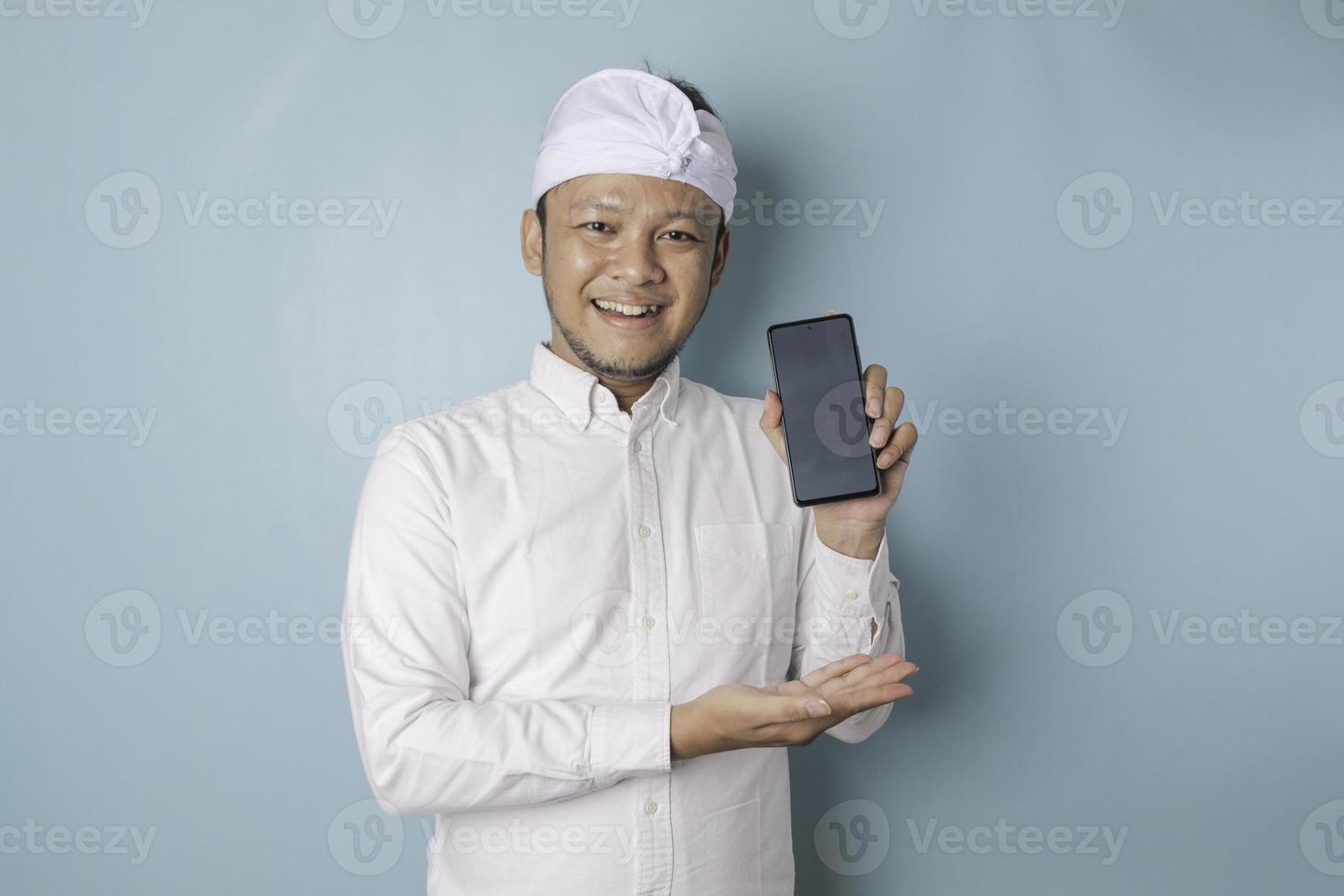 homme balinais excité portant un bandeau traditionnel ou une chemise blanche et udeng pointant vers l'espace de copie sur son smartphone, isolé par fond bleu photo