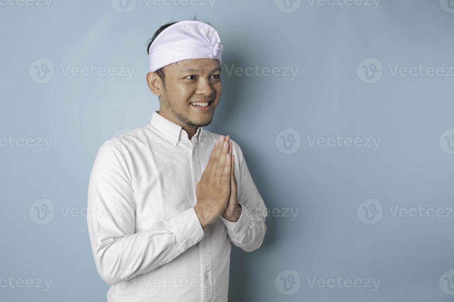 jeune homme balinais souriant portant un bandeau udeng ou traditionnel et une chemise blanche faisant des gestes de salutation ou namaste isolé sur fond bleu photo