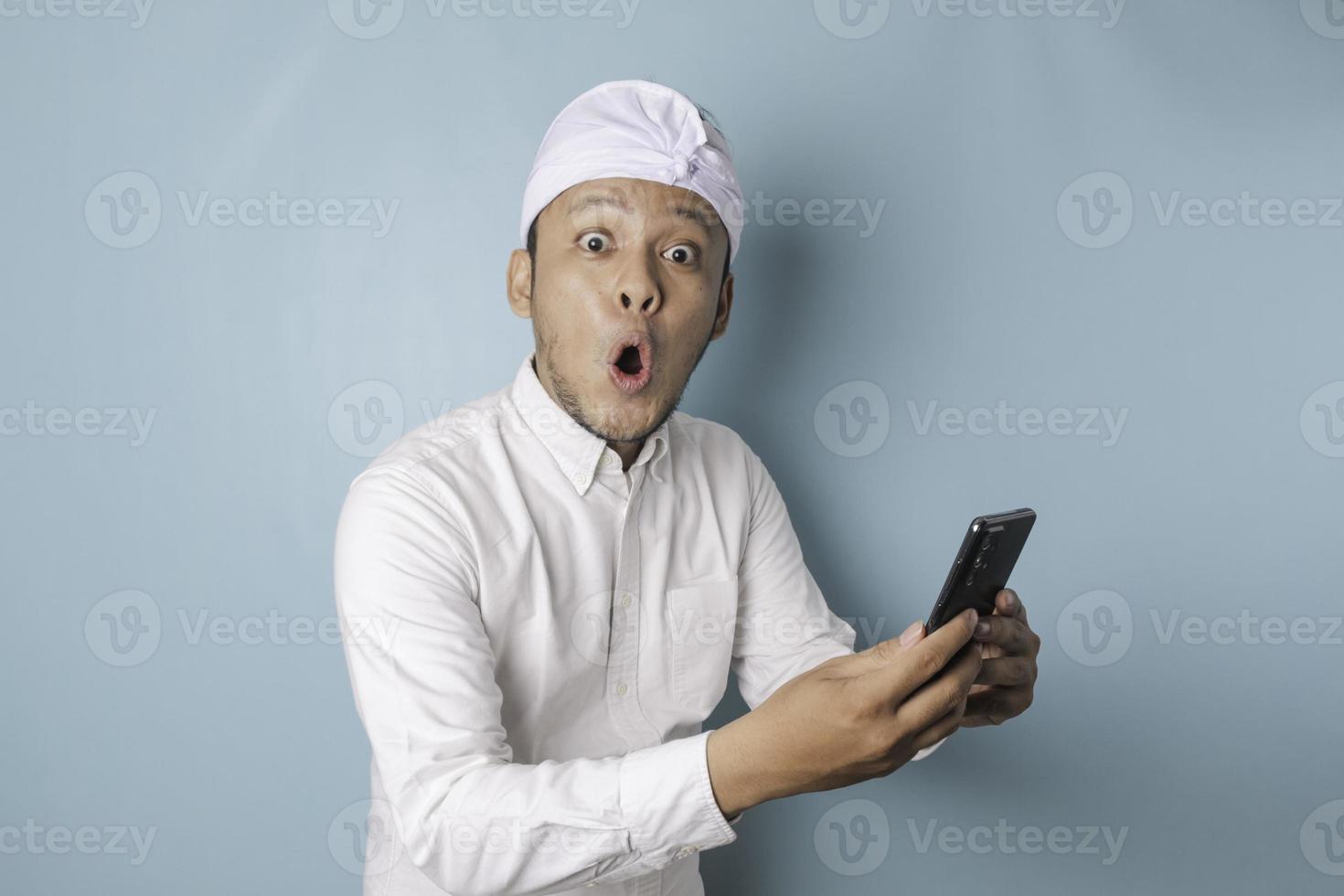 homme balinais surpris portant un bandeau udeng ou traditionnel et une chemise blanche tenant son smartphone, isolé par fond bleu photo
