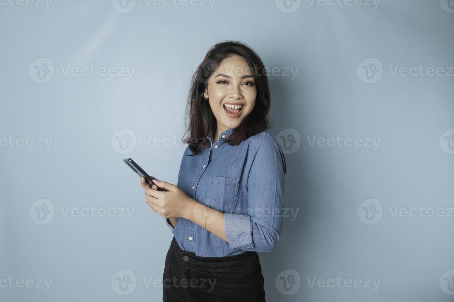 un portrait d'une femme asiatique heureuse sourit et tient son smartphone portant une chemise bleue isolée par un fond bleu photo