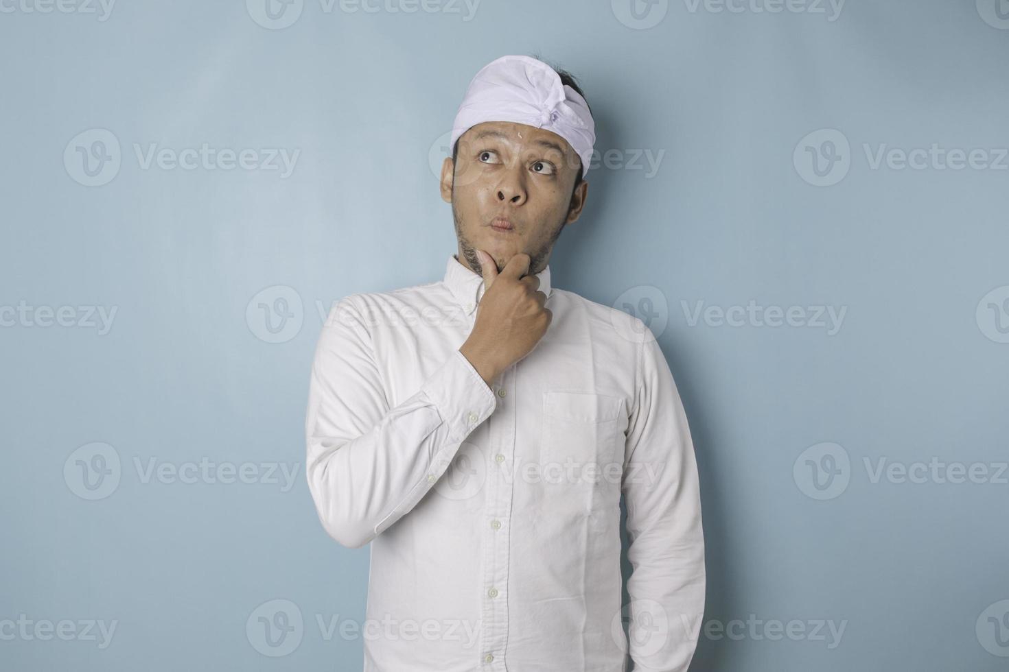 portrait d'un jeune homme balinais réfléchi portant un bandeau udeng ou traditionnel et une chemise blanche regardant de côté tandis que son doigt sur son menton isolé sur fond bleu photo
