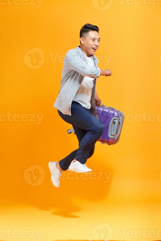 jeune homme asiatique sautant avec sac de voyage valise isolé sur fond jaune photo