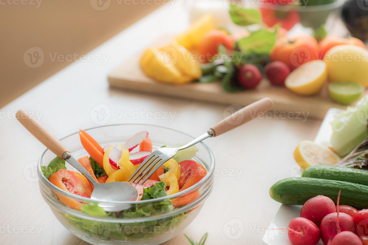 salade de légumes dans un bol en verre transparent et des couverts sur la table de la cuisine, il y a des légumes et des fruits comme les tomates, les concombres, les poivrons, la laitue, les radis et les citrons. photo