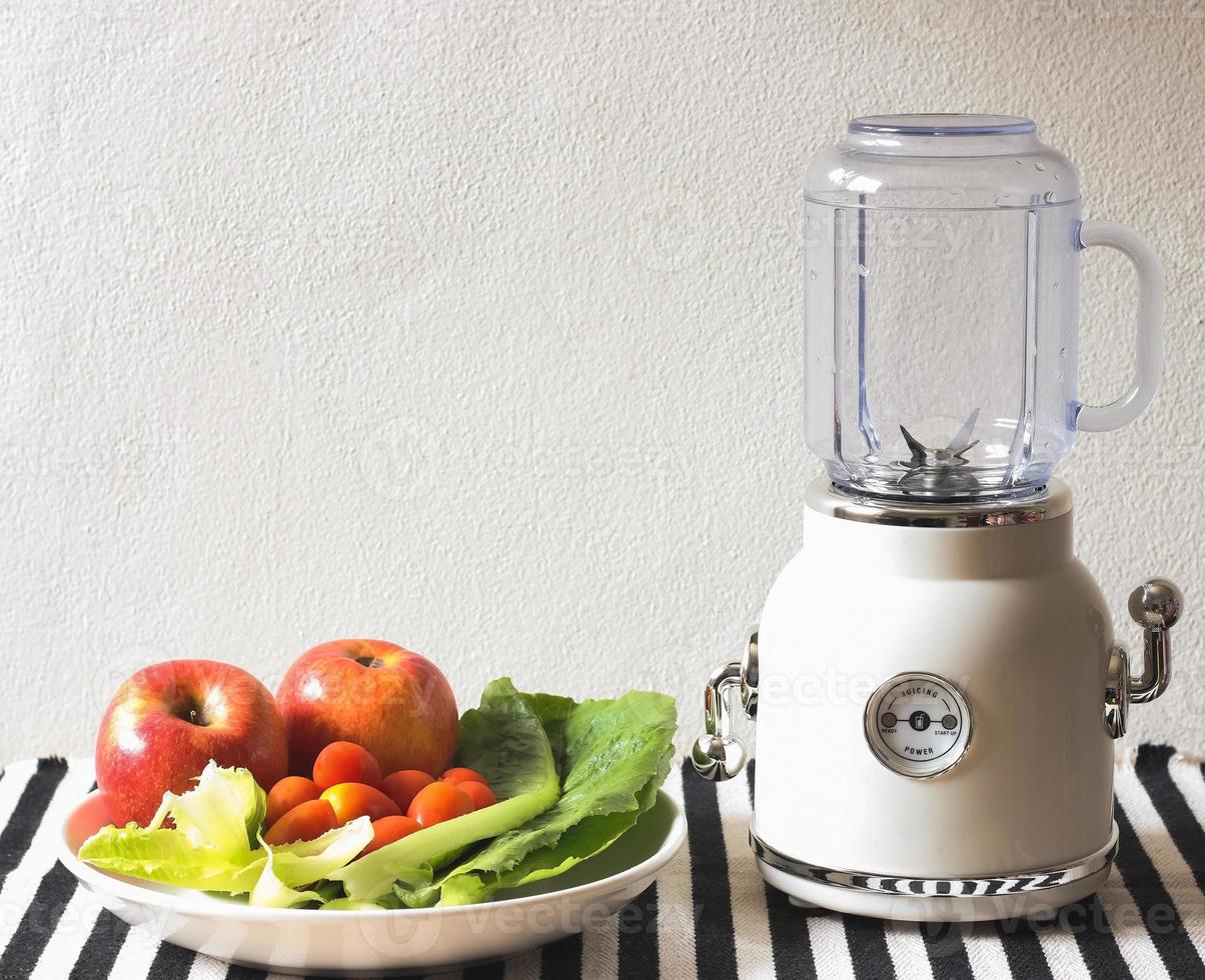 mélangeur vintage blanc vide ou machine à smoothie avec une assiette de légumes, tomates et pommes sur une nappe à rayures noires et blanches et un mur blanc. préparation pour la fabrication de smoothies. photo