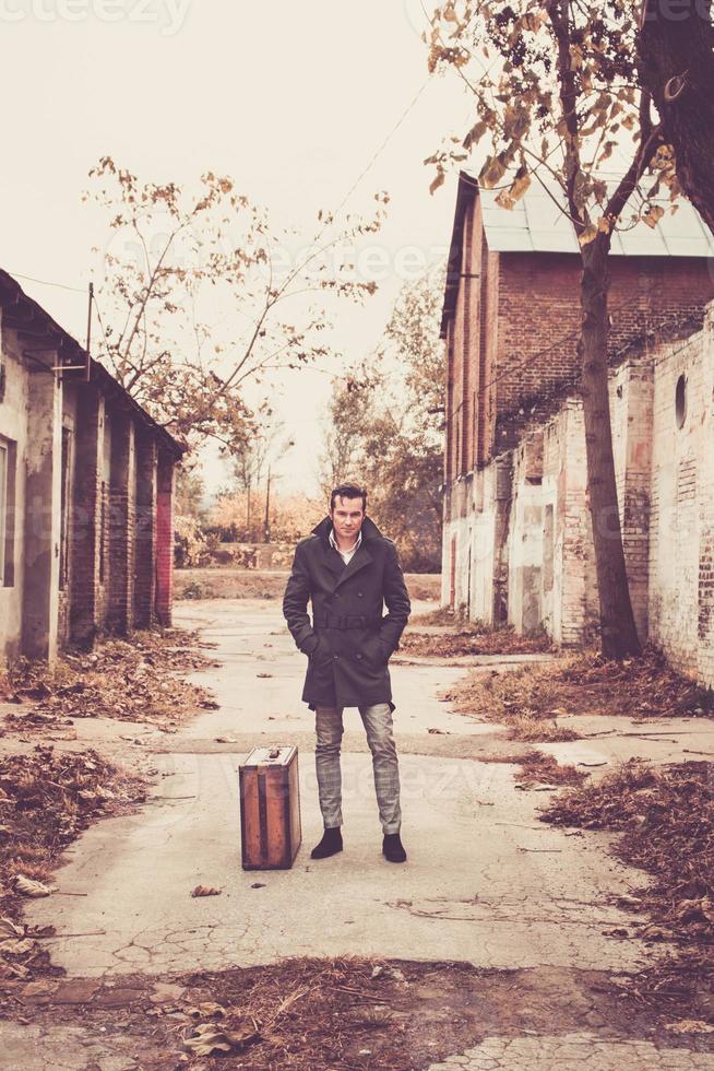 homme en manteau avec valise vintage debout au milieu de bâtiments en ruine. photo