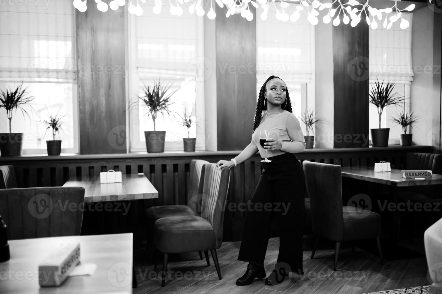 portrait d'une jeune femme afro-américaine séduisante portant un pull vert et un jean noir posent au restaurant avec un verre de vin rouge à portée de main. photo