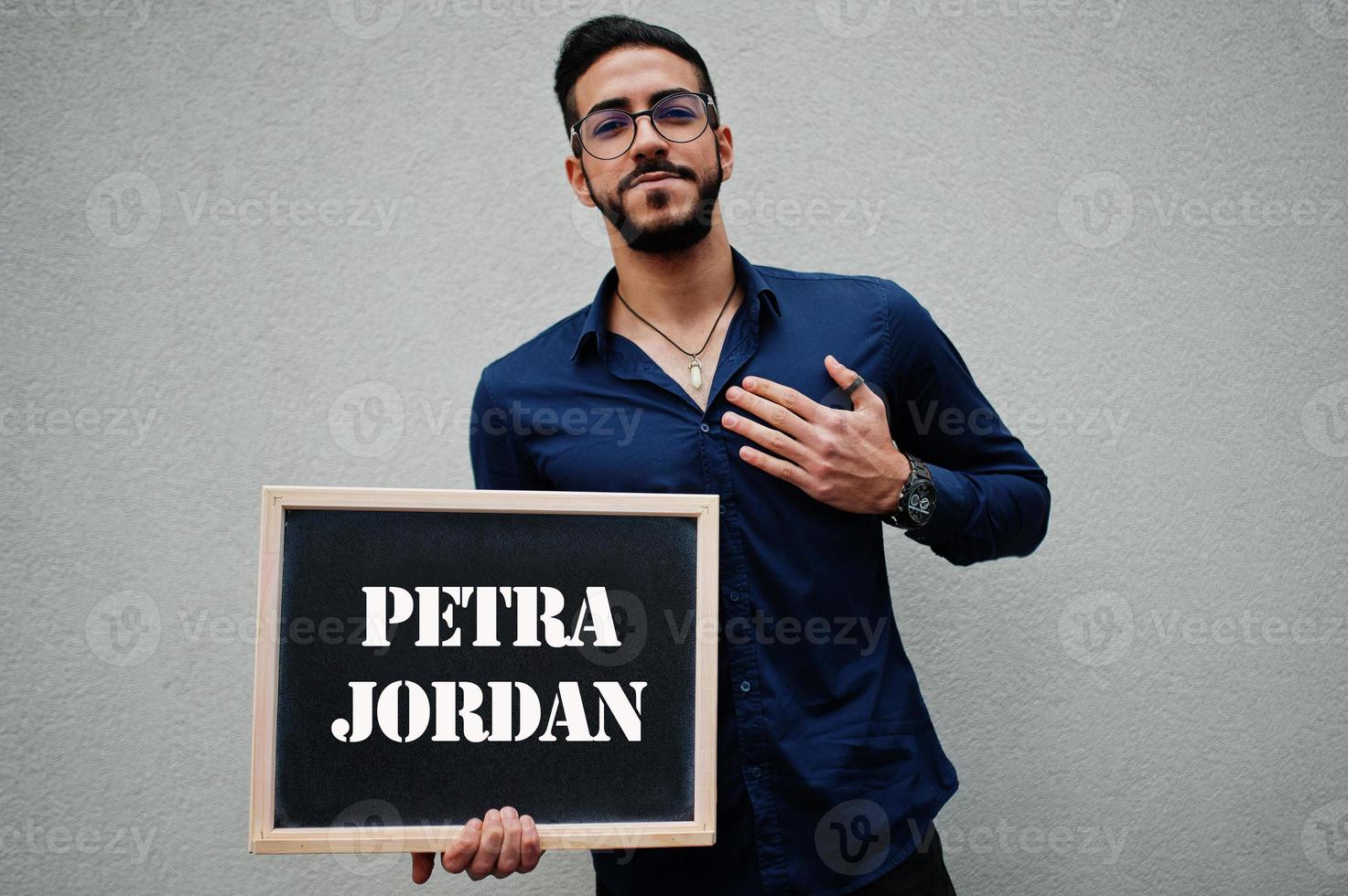 l'homme arabe porte une chemise bleue et des lunettes tient le tableau avec l'inscription de petra jordan. plus grandes villes du concept du monde islamique. photo