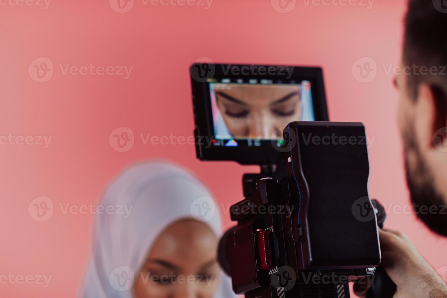 vidéaste en studio numérique enregistrant une vidéo sur une caméra professionnelle en tirant sur une femme musulmane portant une écharpe hijab sur fond rose en plastique. photo