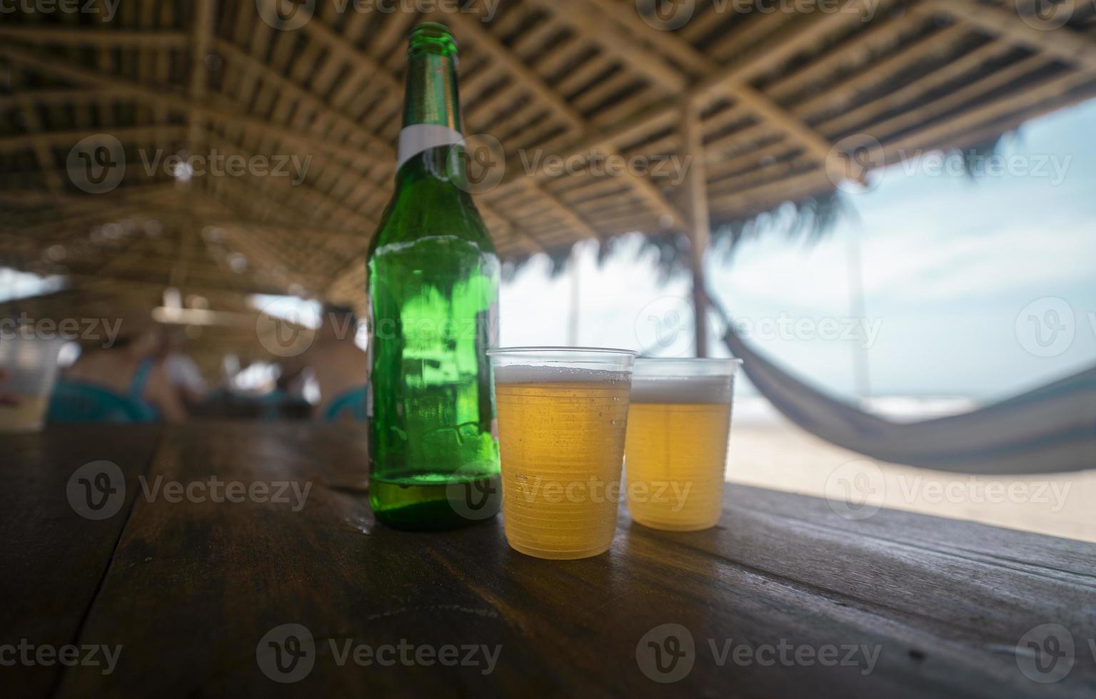 Deux verres en plastique avec de la bière et une bouteille verte sur une table en bois dans un restaurant sur la plage avec un toit en bambou et des hamacs sur les côtés photo