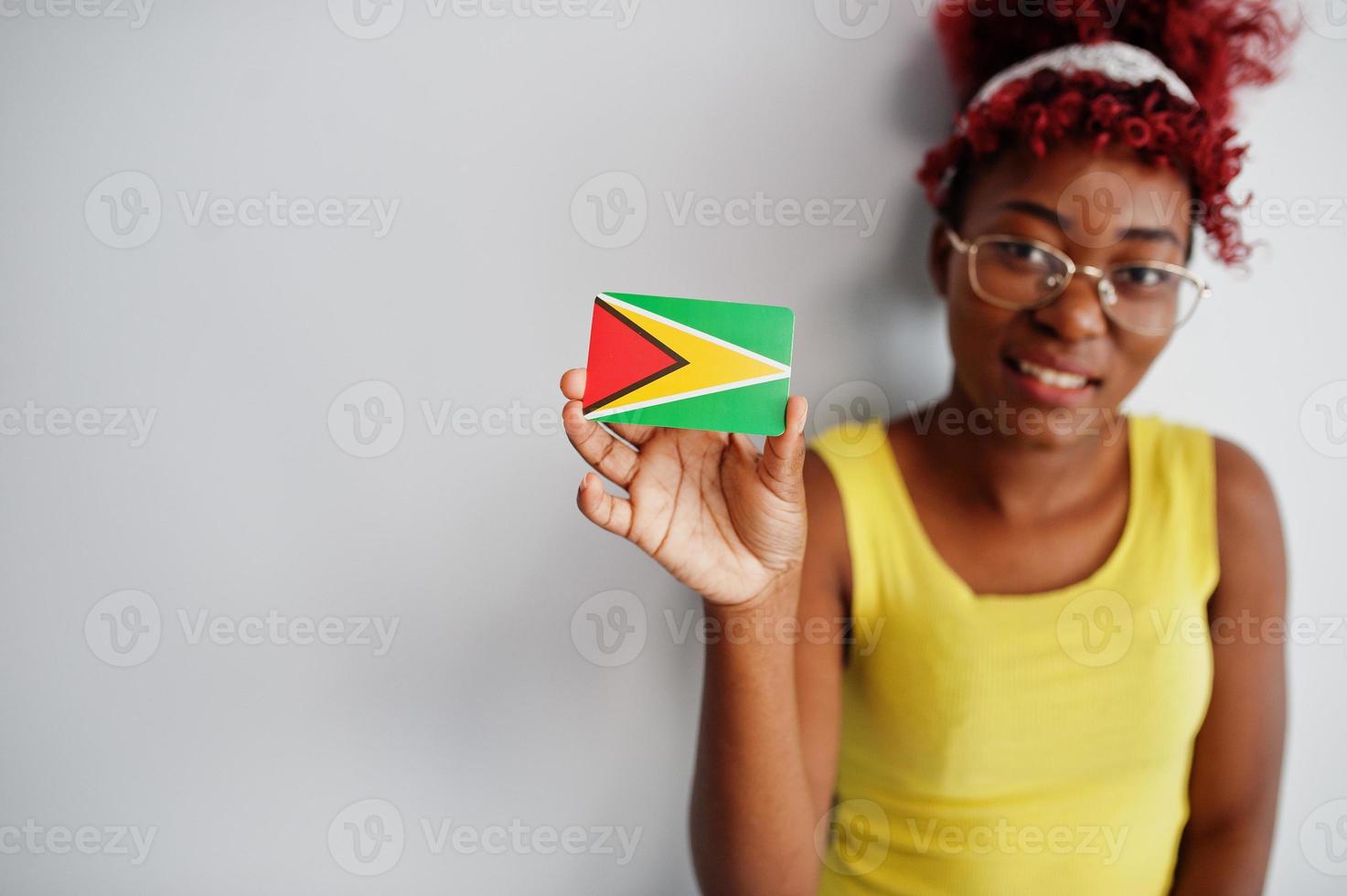 femme afro-américaine aux cheveux afro, porter un singulet jaune et des lunettes, tenir le drapeau de la guyane isolé sur fond blanc. photo