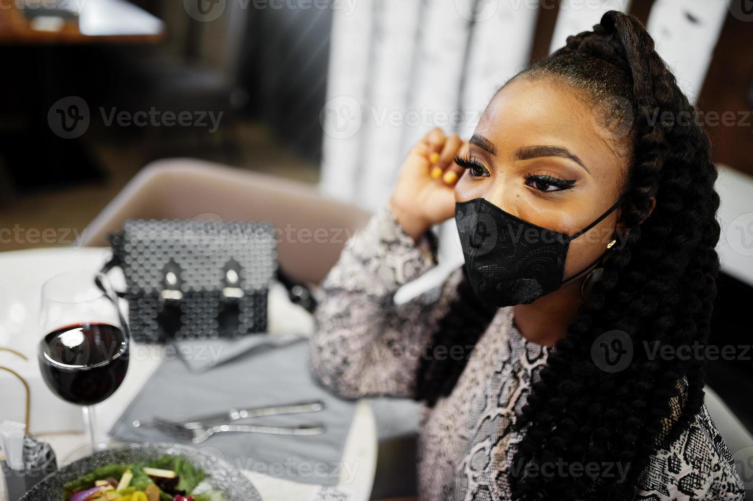 jeune femme afro-américaine élégante, portant un masque noir, assise au restaurant, dégustant une nourriture saine avec du vin. photo