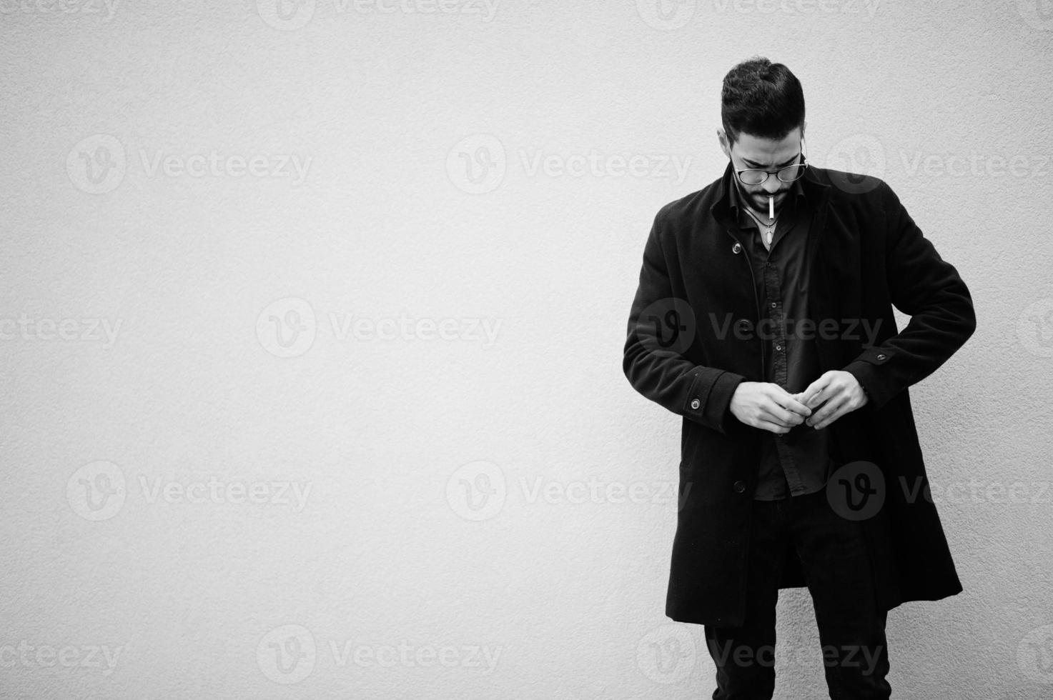 un entrepreneur du moyen-orient porte un manteau noir et une chemise bleue, des lunettes debout contre un mur blanc et fume une cigarette. photo