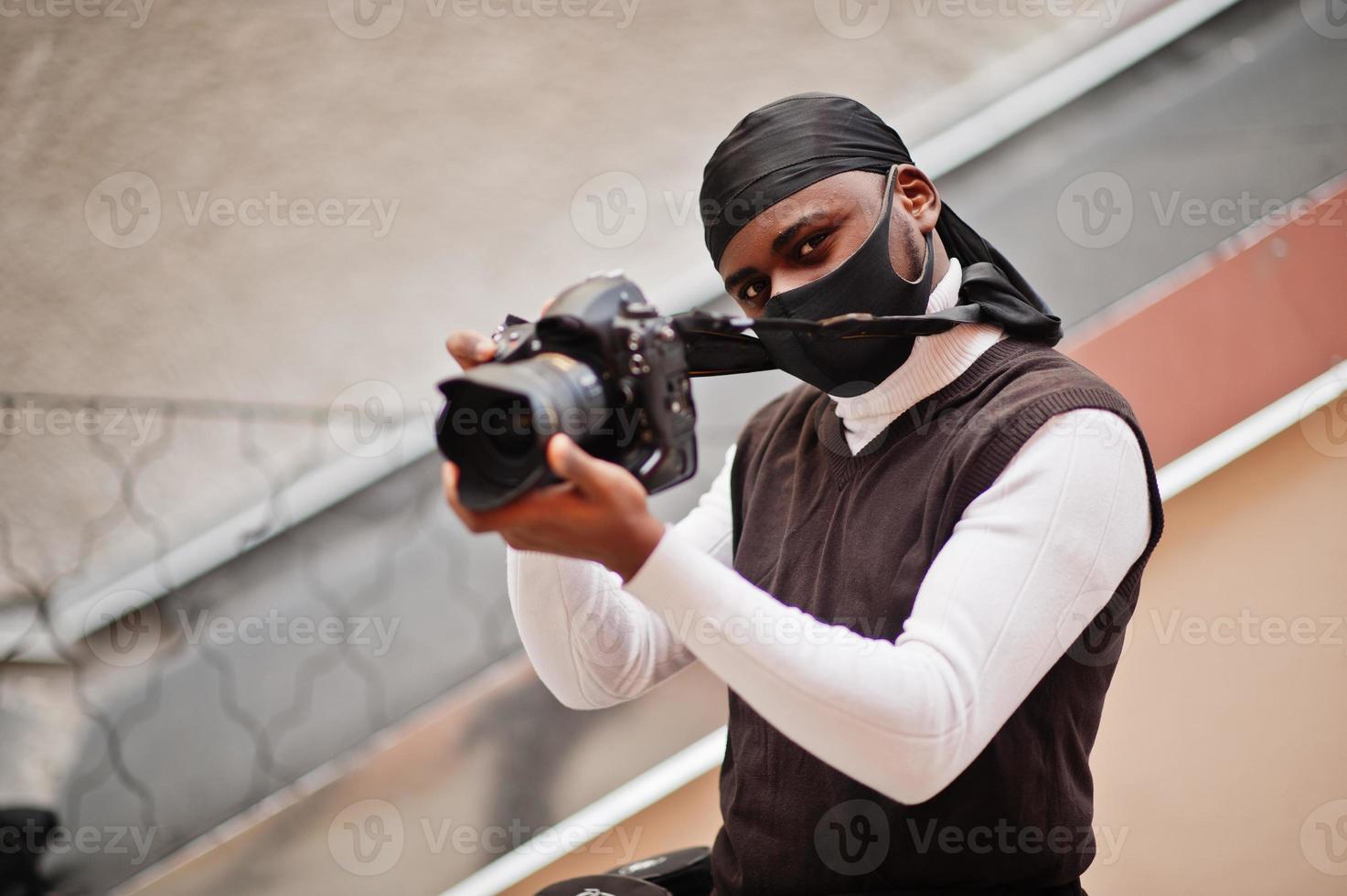 jeune vidéaste afro-américain professionnel tenant une caméra professionnelle avec un équipement professionnel. caméraman afro portant du duraq noir et un masque de protection du visage, faisant des vidéos. photo