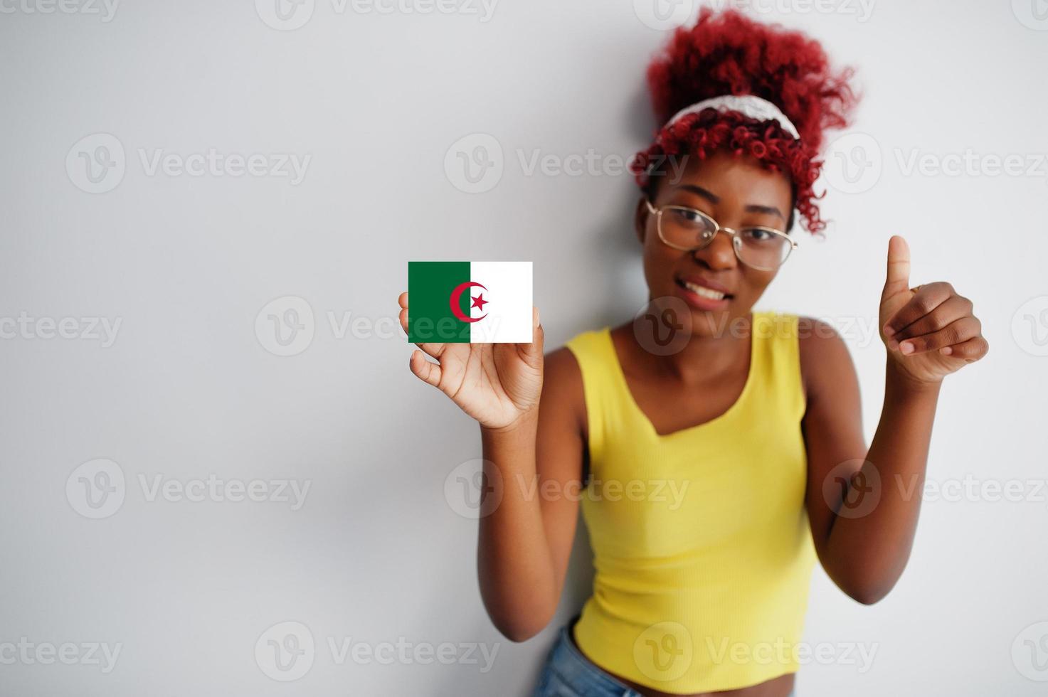 femme africaine aux cheveux afro, porter un maillot jaune et des lunettes, tenir le drapeau algérien isolé sur fond blanc, montrer le pouce vers le haut. photo