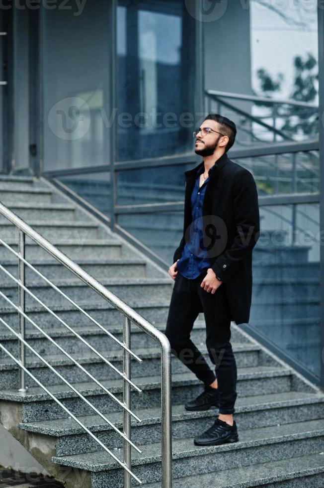 un entrepreneur du moyen-orient porte un manteau noir et une chemise bleue, des lunettes contre un immeuble de bureaux, se tient dans les escaliers. photo