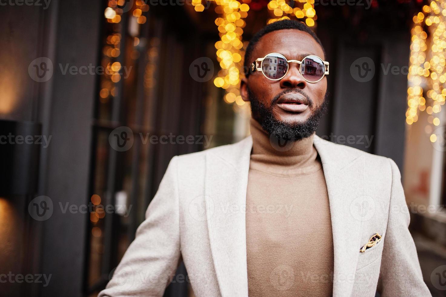 un homme afro-américain élégant porte une veste beige avec des lunettes de soleil pose contre la maison avec une guirlande dorée. photo