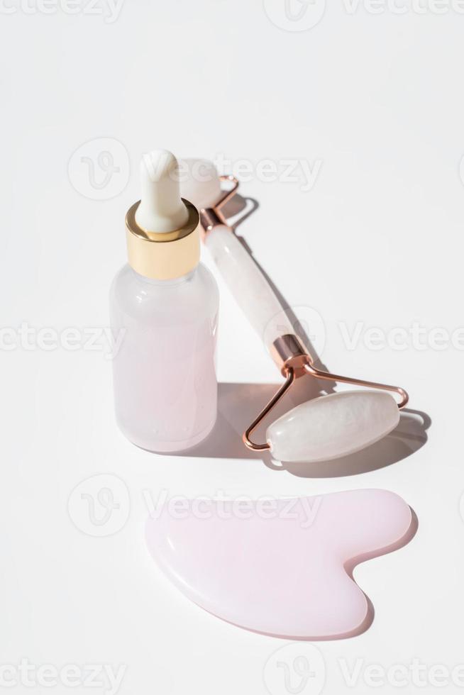 rouleau facial, gua sha et sérum hydratant sur fond blanc. cosmétiques naturels et outils pour le visage. rouleau de beauté en quartz rose, outils heart gua sha pour les routines de soins de la peau. ombres dures photo