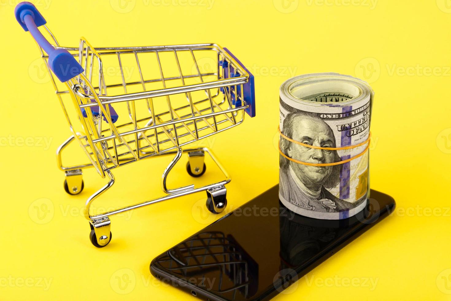 téléphone portable avec un rouleau de billets de cent dollars et un panier sur fond jaune. concept d'achat en ligne. espace de copie. photo
