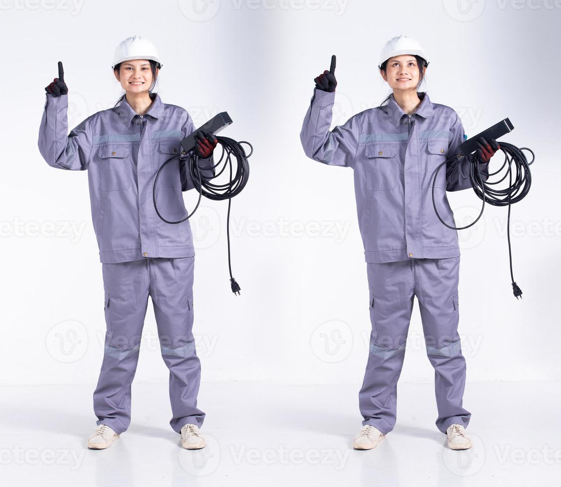 pleine longueur 20s jeune femme électricienne ingénieur de course mixte, pointant le doigt vers le haut dans l'air, porter un uniforme gris. Femme entrepreneur tenir le câble de prise électrique sourire heureux sur fond blanc isolé photo