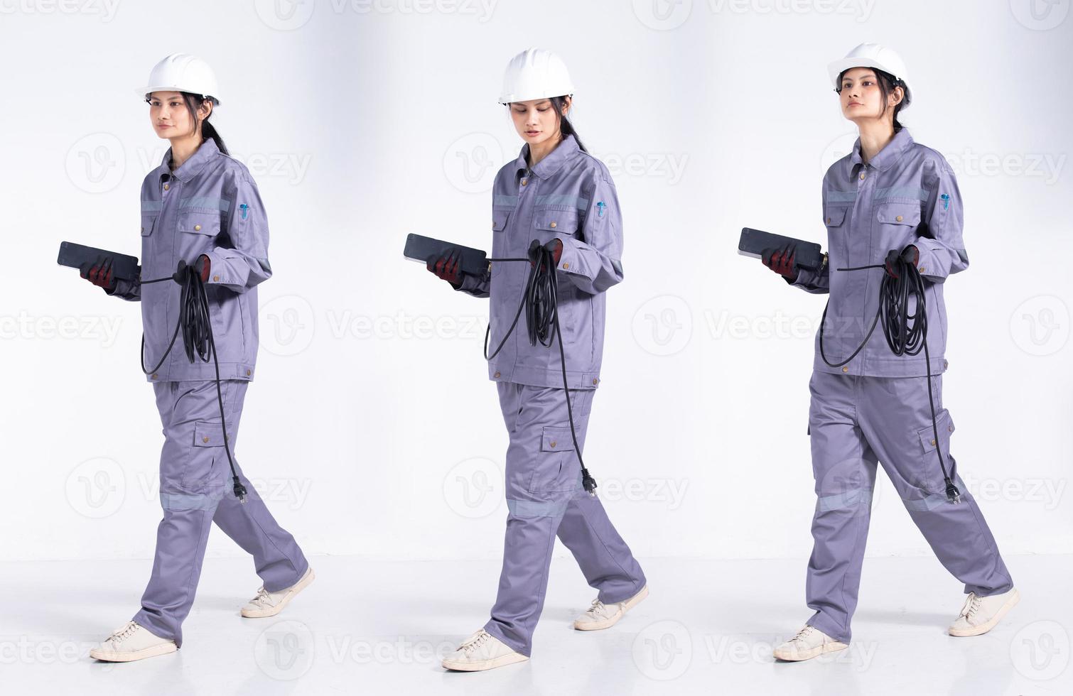 pleine longueur 20s jeune ingénieur de course mixte femme électricienne, marchant vers l'avant gauche droite, porter un uniforme gris. Femme entrepreneur tenir le câble de prise électrique sourire heureux sur fond blanc isolé photo