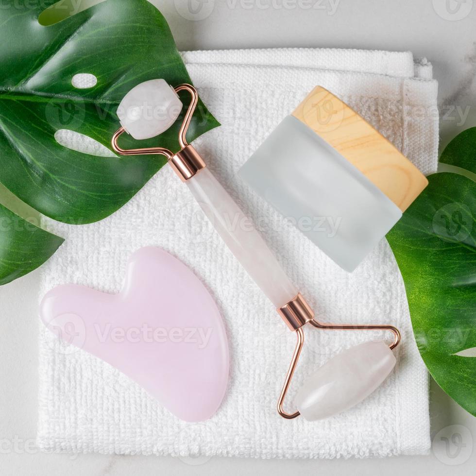 kit facial pour les soins de la peau à domicile et le spa. rouleau de quartz rose, masseurs gua sha et pot de crème pour le visage sur fond de serviette blanche avec feuilles de monstara vertes. concept de traitement naturel. vue de dessus. photo