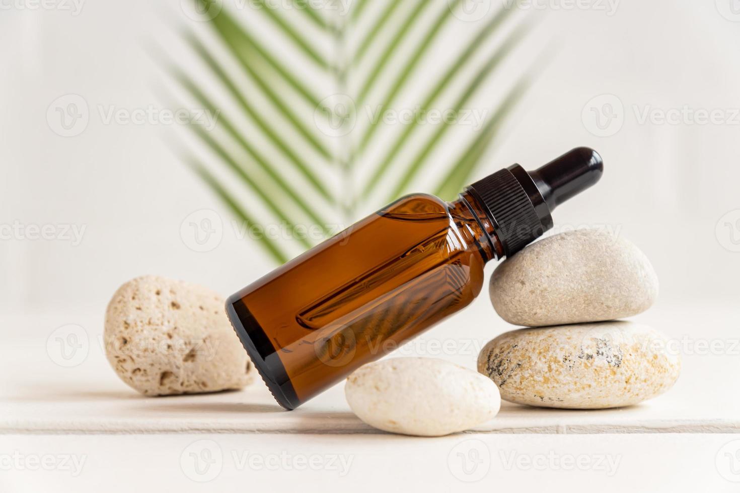bouteille de verre ambré foncé avec huile essentielle ou sérum pour le visage sur pile de pierres naturelles sur fond de feuilles tropicales. photo