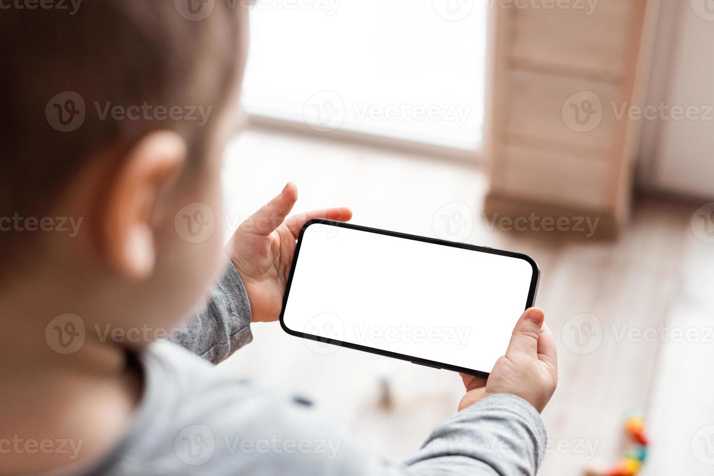 les mains du tout-petit garçon tenant le téléphone sur le fond de la maison. les mains de l'enfant tiennent un smartphone noir et regardent une vidéo en position horizontale. écran vide. fermer. photo