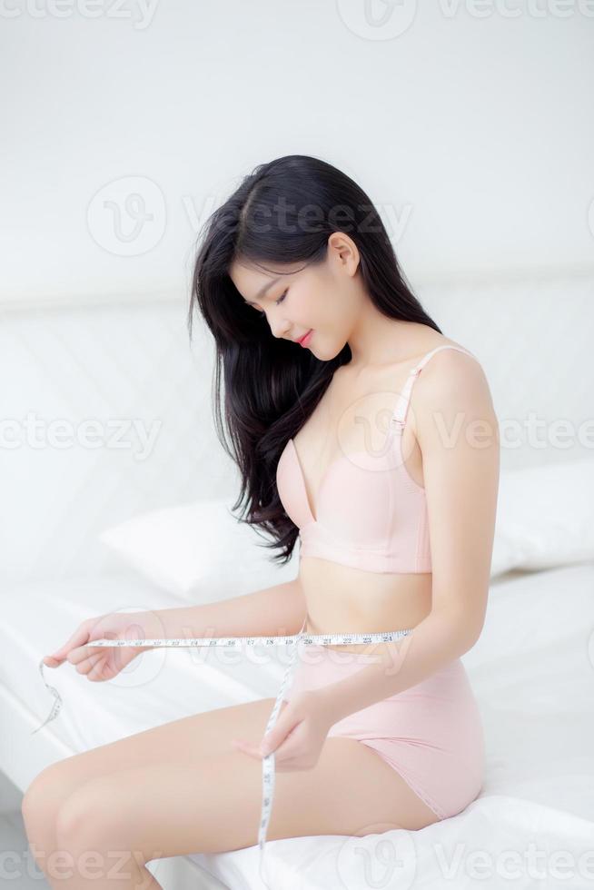 belle jeune femme asiatique corps sexy mince mesurant l'abdomen pour contrôler la perte de poids dans la chambre, belle fille ventre mince ont de la cellulite avec un ruban à mesurer pour le concept de régime, de santé et de bien-être. photo