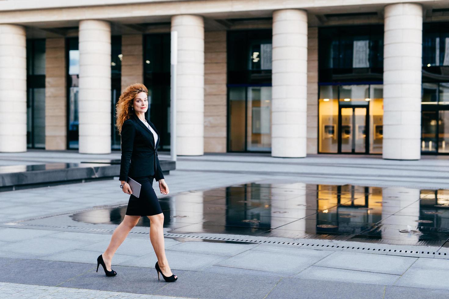 élégante femme mince en costume noir et chaussures à talons hauts, tenant la tablette dans les mains pour aller travailler. femme d'affaires en mouvement. belle jeune femme confiante en vêtements formels près de l'immeuble de bureaux photo