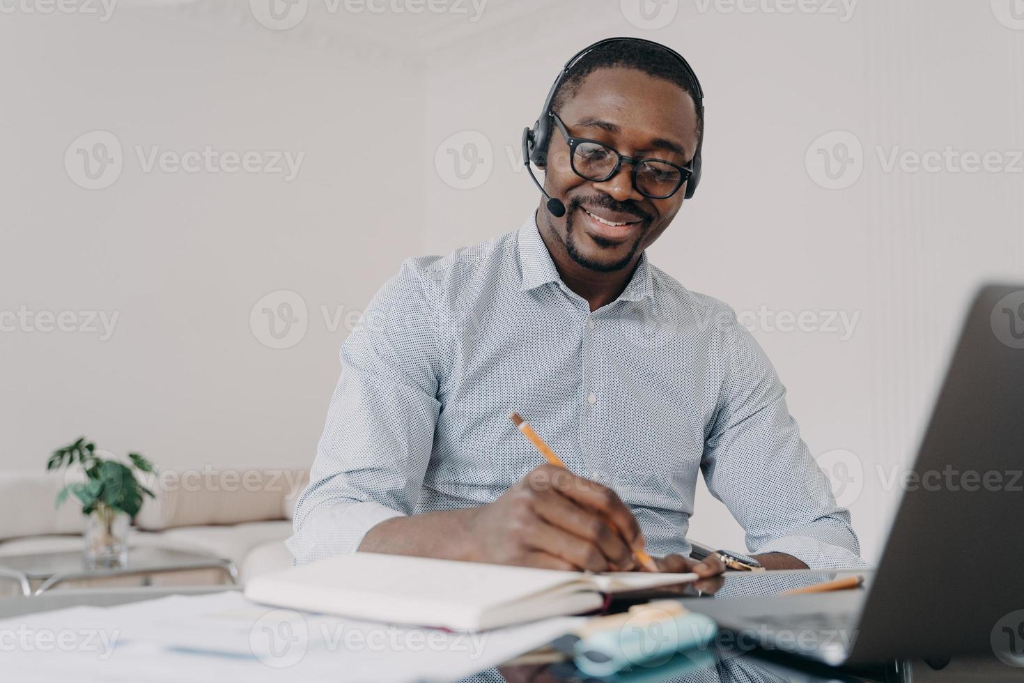 un homme afro-américain dans un casque apprend en ligne sur un ordinateur portable, prend des notes. e-learning, enseignement à distance photo