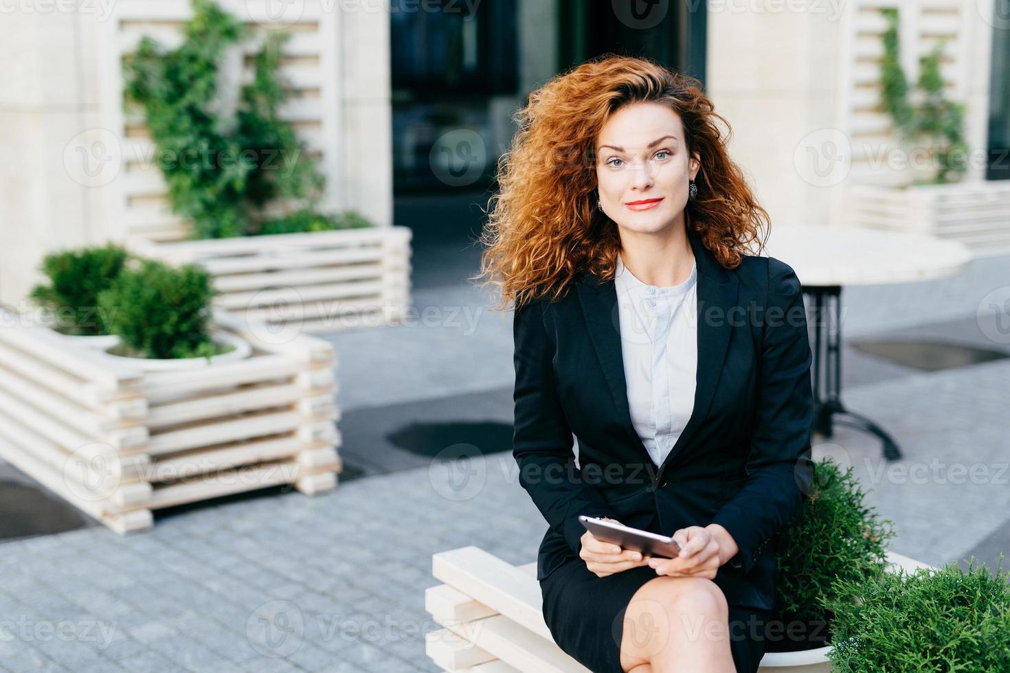 belle femme magnifique aux cheveux touffus ondulés, vêtue d'un costume formel, assise les jambes croisées au café en plein air, tenant une tablette en attendant quelqu'un, regardant la caméra avec un regard confiant photo