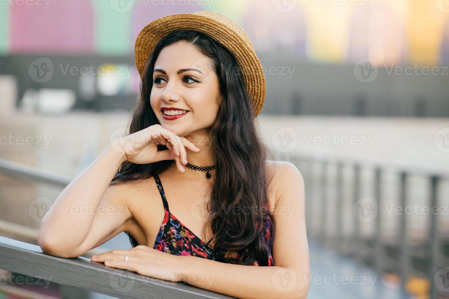 jeune modèle féminin avec de longs cheveux noirs et épais debout au pont ayant une expression rêveuse regardant au loin, portant un chapeau d'été et une robe se reposant à l'extérieur. personnes, détente, concept de beauté photo